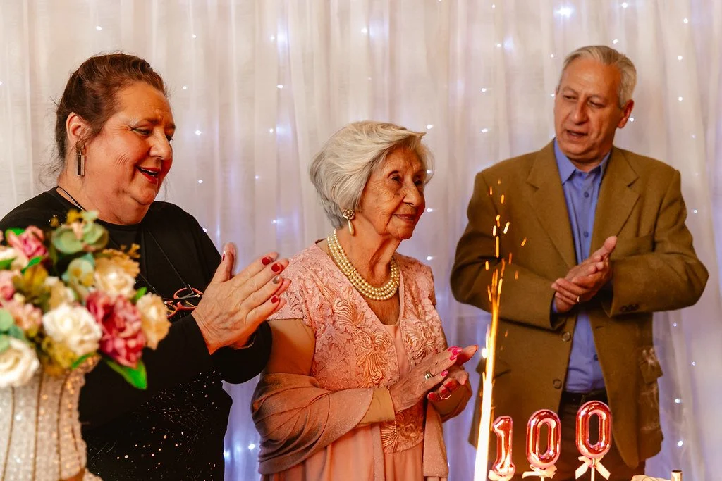 Idosa comemorando seu 100º aniversário com duas outras pessoas, uma mulher e um homem, em uma festa com decoração de luzes de fadas. Há um bolo com o número 100 na foto.