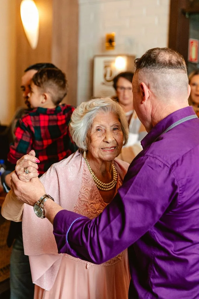 Um idoso e um homem dançam juntos em um ambiente interno de restaurante ou sala de jantar, com outras pessoas ao fundo, incluindo uma criança e duas mulheres. Ambos parecem felizes e a cena transmite um momento de celebração ou confraternização.