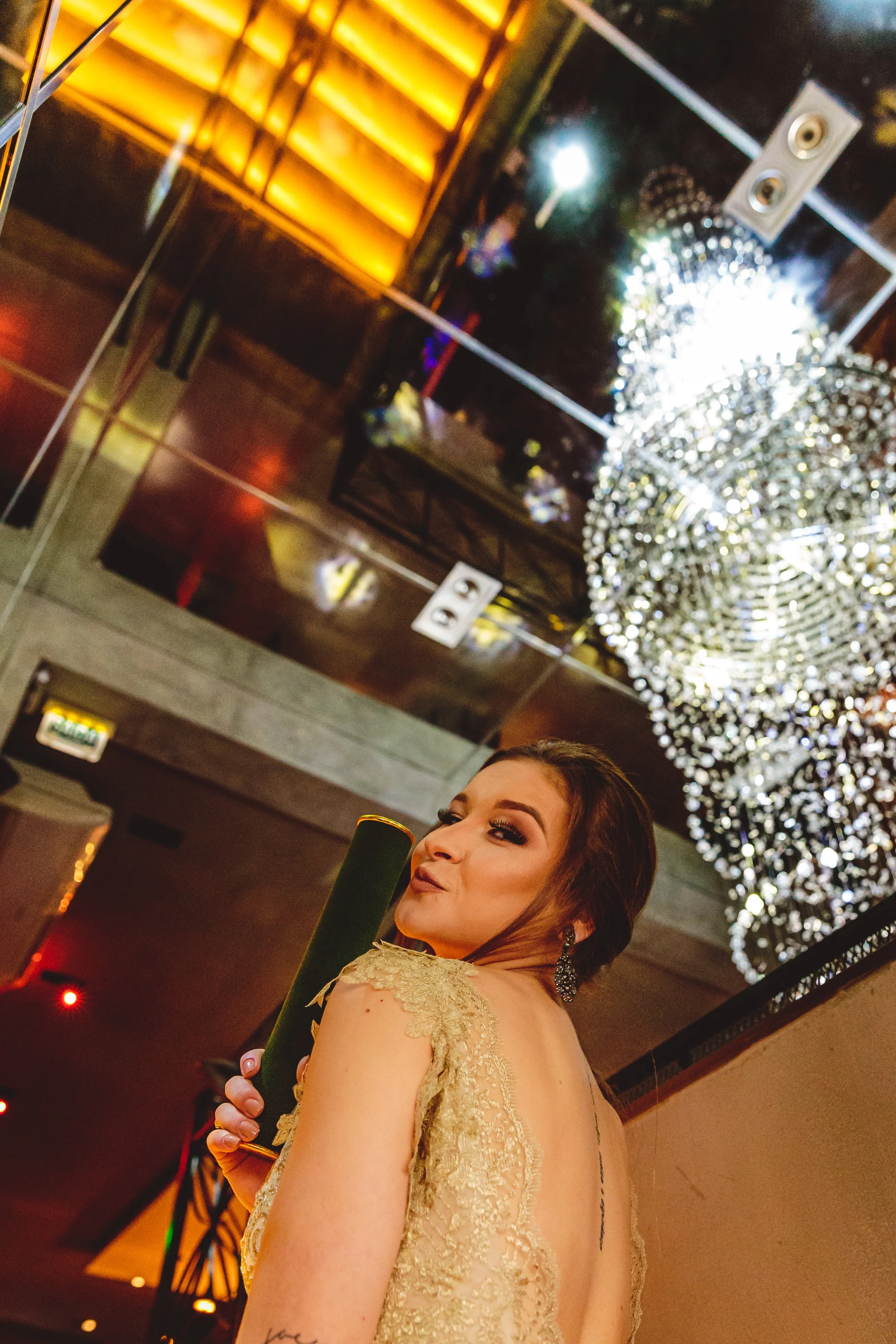 Mulher em vestido dourado, segurando um presente, em evento elegante com um lustre de cristal no teto.