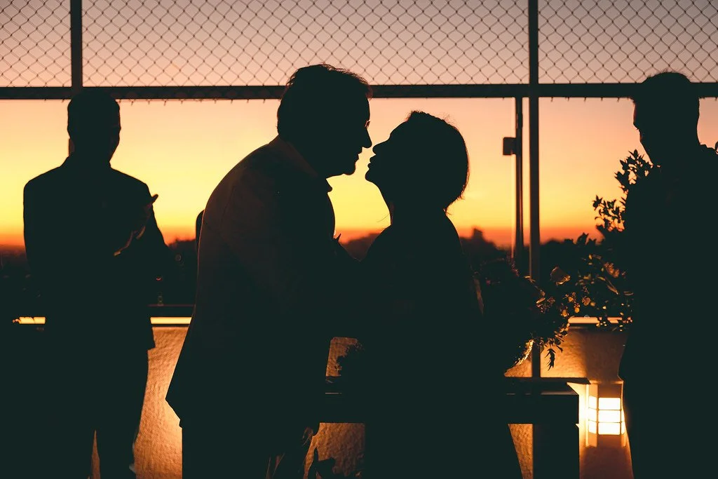 Silhueta de um casal se beijando ao pôr do sol, com dois homens ao fundo em um ambiente ao ar livre.