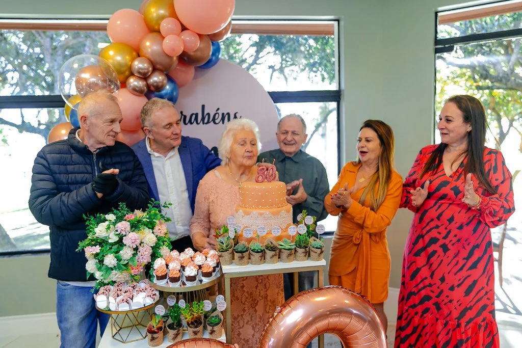 Grupo de pessoas comemorando o aniversário de uma idosa, com uma mesa de bolo decorada, cupcakes e plantas, fundo com balões coloridos e uma placa de parabéns.