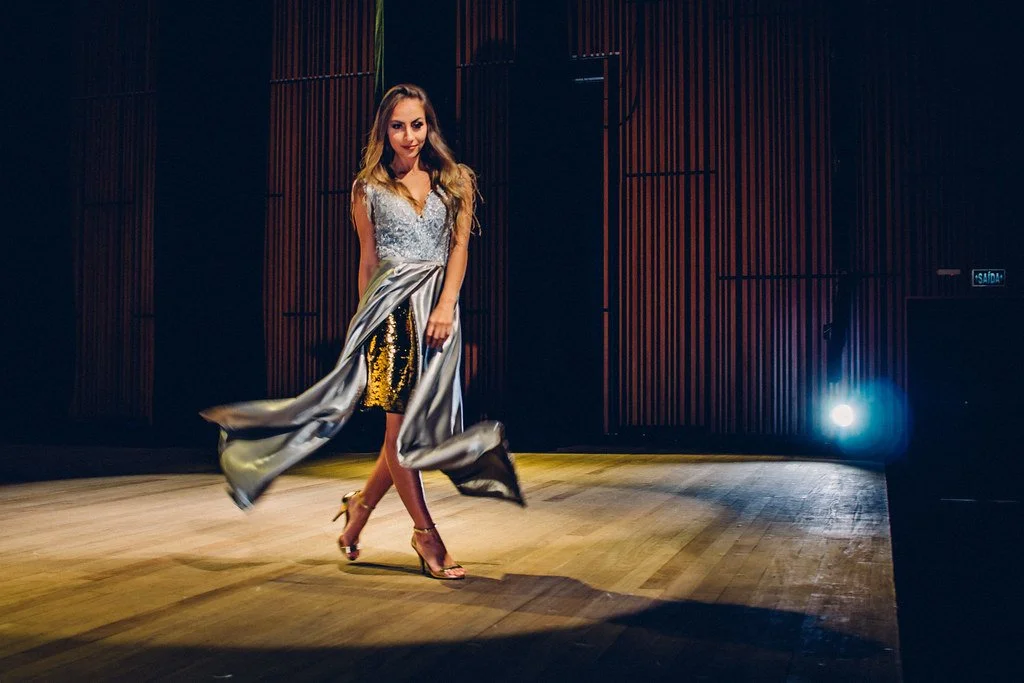 Mulher vestida com roupa de gala, caminhando no palco com iluminação dramática ao fundo.
