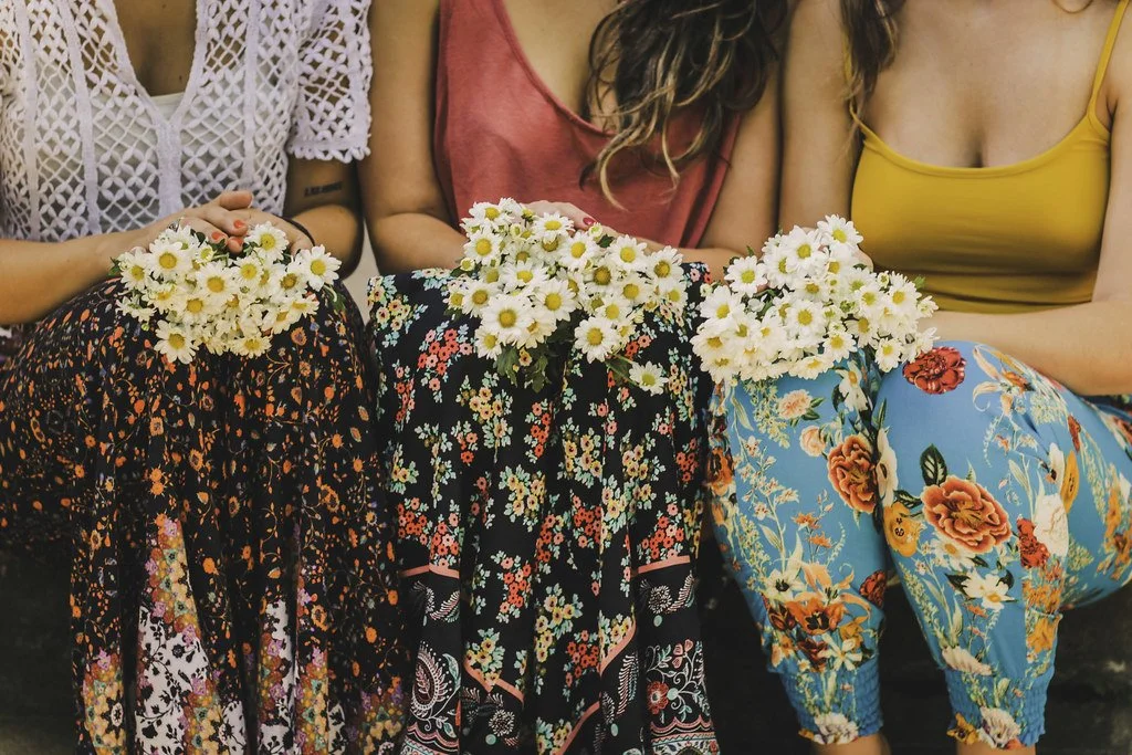 Três mulheres sentadas com flores de campo na mão, usando vestidos florais coloridos.