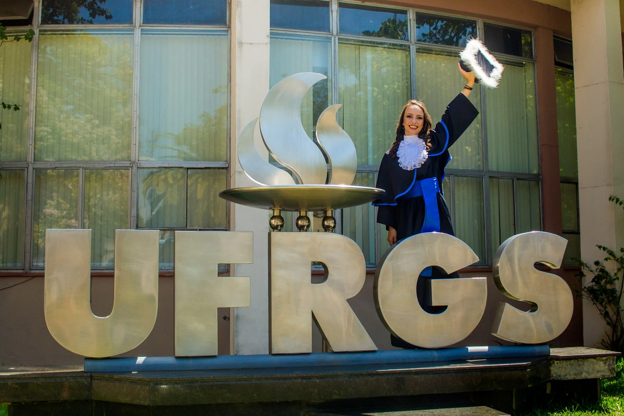 Pessoa em traje de formatura segurando chapéu de formatura ao lado de uma escultura de letras grandes que formam a palavra "UERGS" e uma chama metálica ao centro, em frente a uma edificação com janelas.