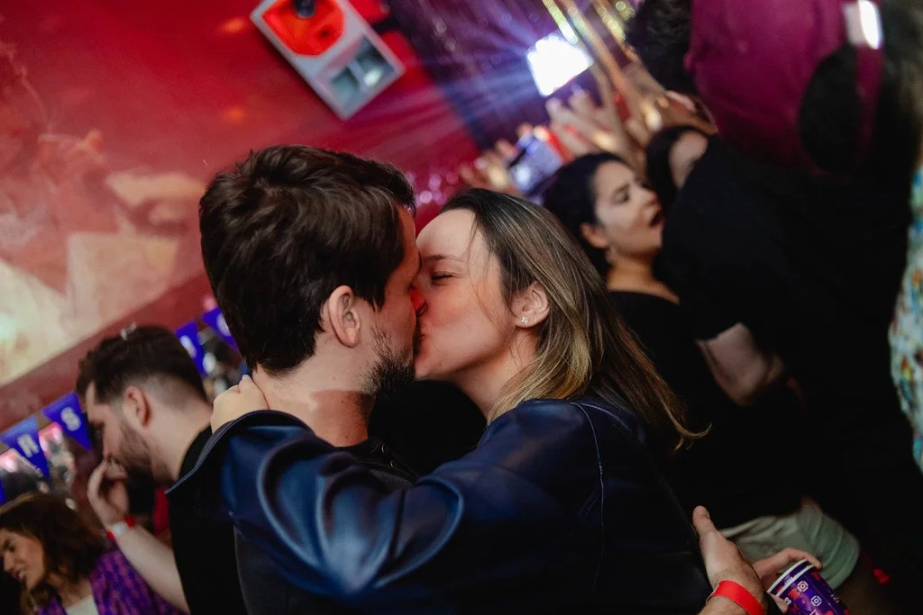 Casal se beijando em um ambiente de festa ou bar com outras pessoas ao fundo.