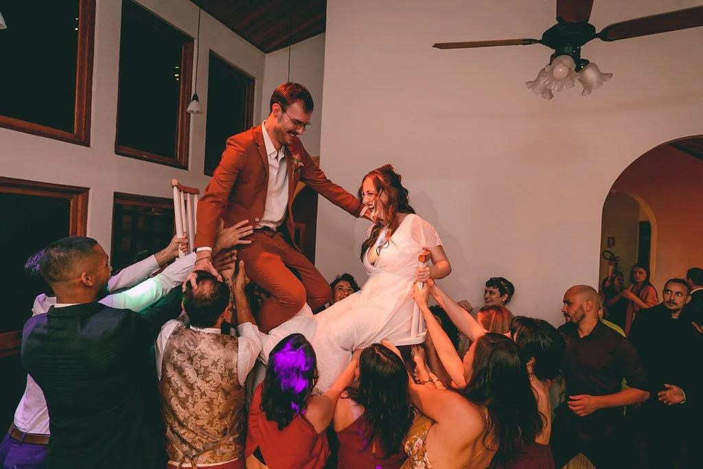 Casal de noivos sendo carregado por amigos durante casamento em ambiente interno.