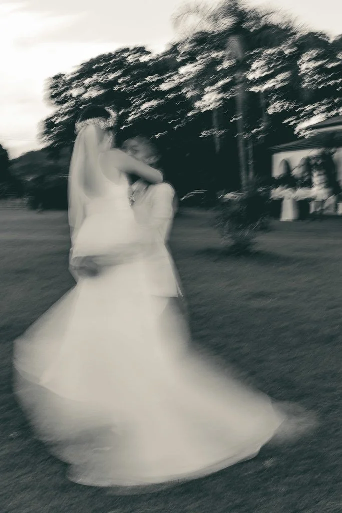 Casal de noivos dançando ao ar livre, com árvores ao fundo, em movimento e borrado na foto.