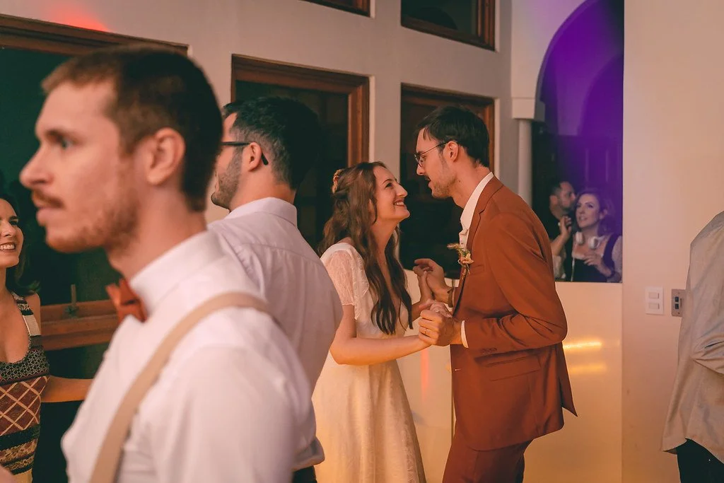 Casal dançando em festa, sorrindo e olhando um ao outro, com várias pessoas ao fundo.