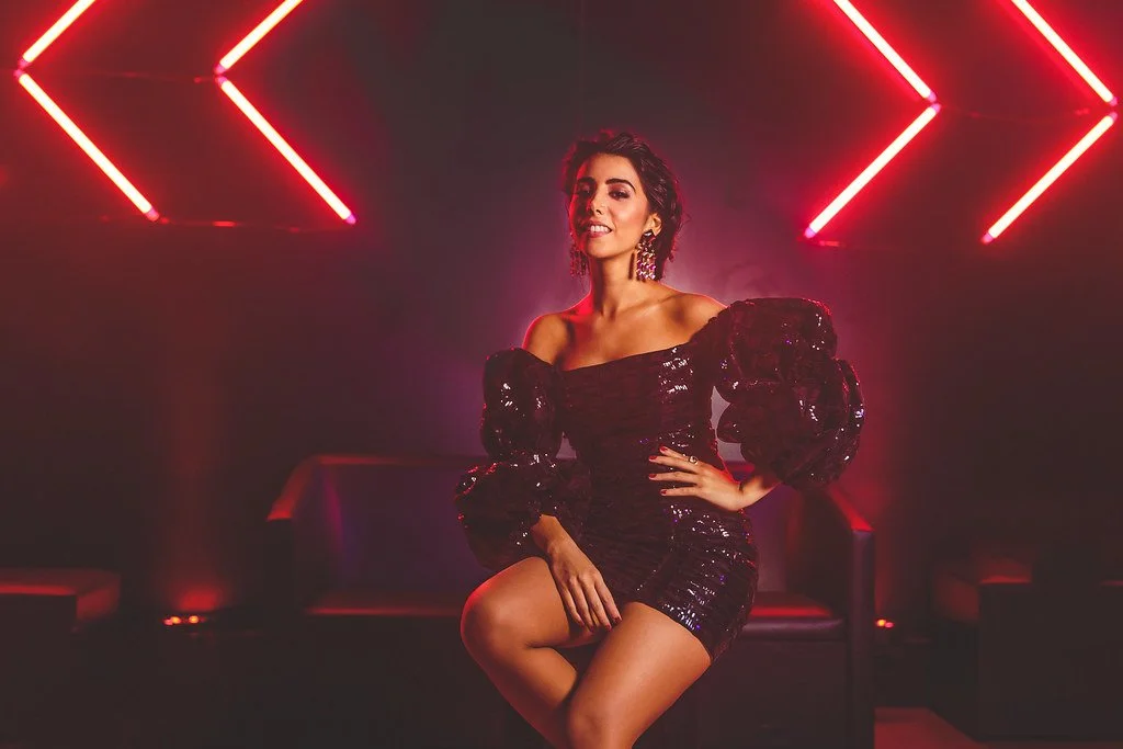 Mulher sentada em ambiente com iluminação neon vermelha, usando vestido preto com brilho e brincos longos, posando com confiança.