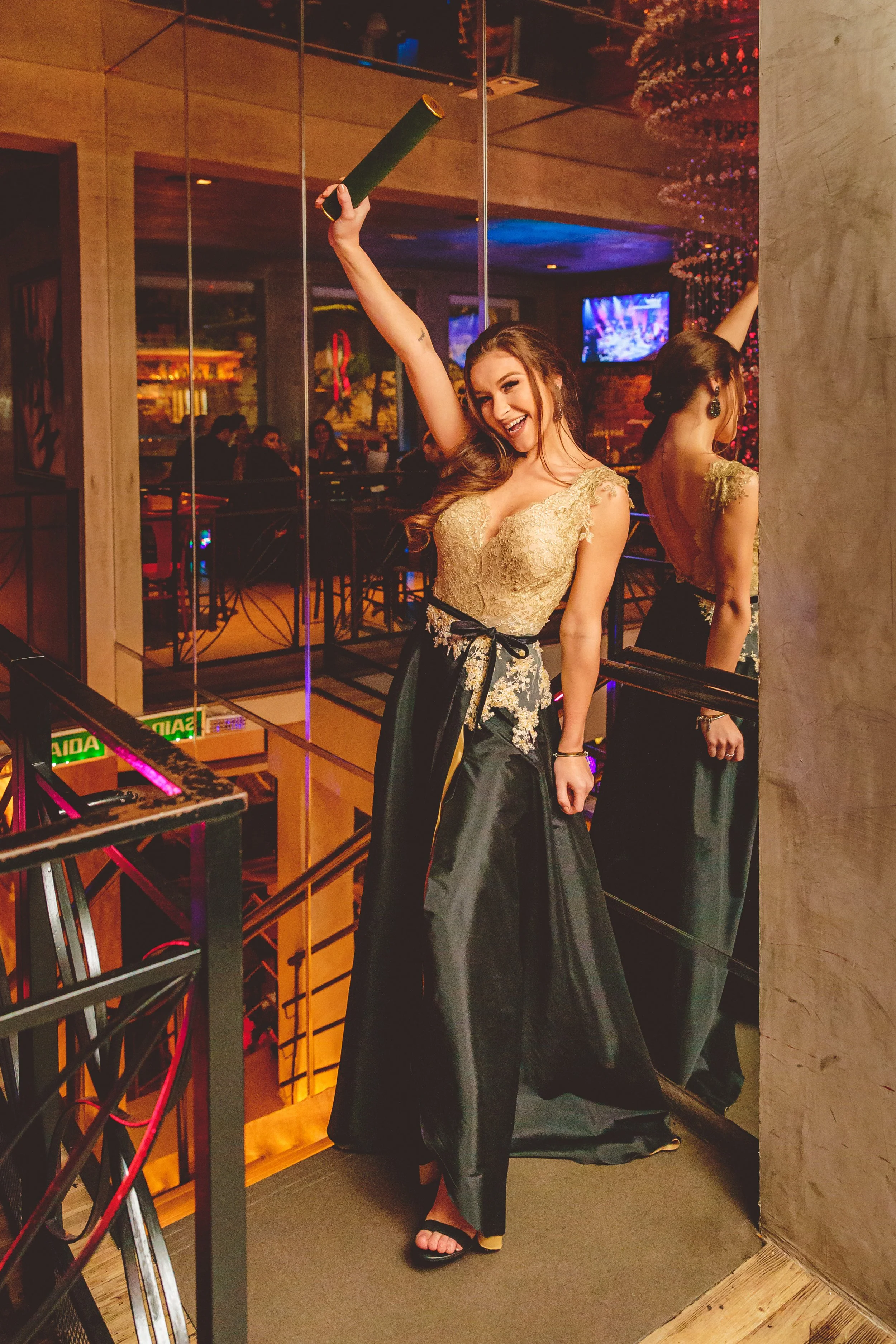 Mulher sorridente em um vestido formal, posando na escadaria de um restaurante ou bar com decoração moderna e iluminação colorida.