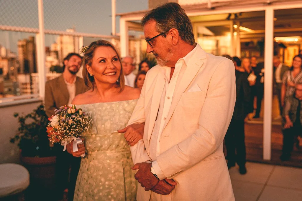 Casal sorridente em um evento, a mulher segura um buquê de flores, ambos vestindo roupas elegantes, com pessoas ao fundo em um ambiente ao ar livre durante o pôr do sol.