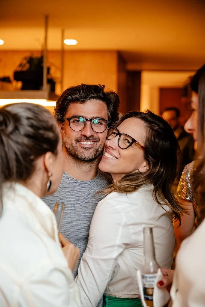 Pessoas sorrindo e brindando em uma festa, com fundo de ambiente interno com iluminação quente.