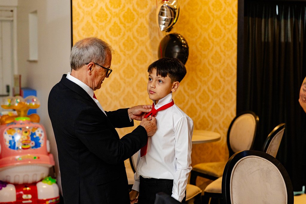 Um menino de terno branco e gravata vermelha sendo presenteado com uma gravata, por um homem mais velho em um ambiente decorado com balões e papel de parede amarelo.