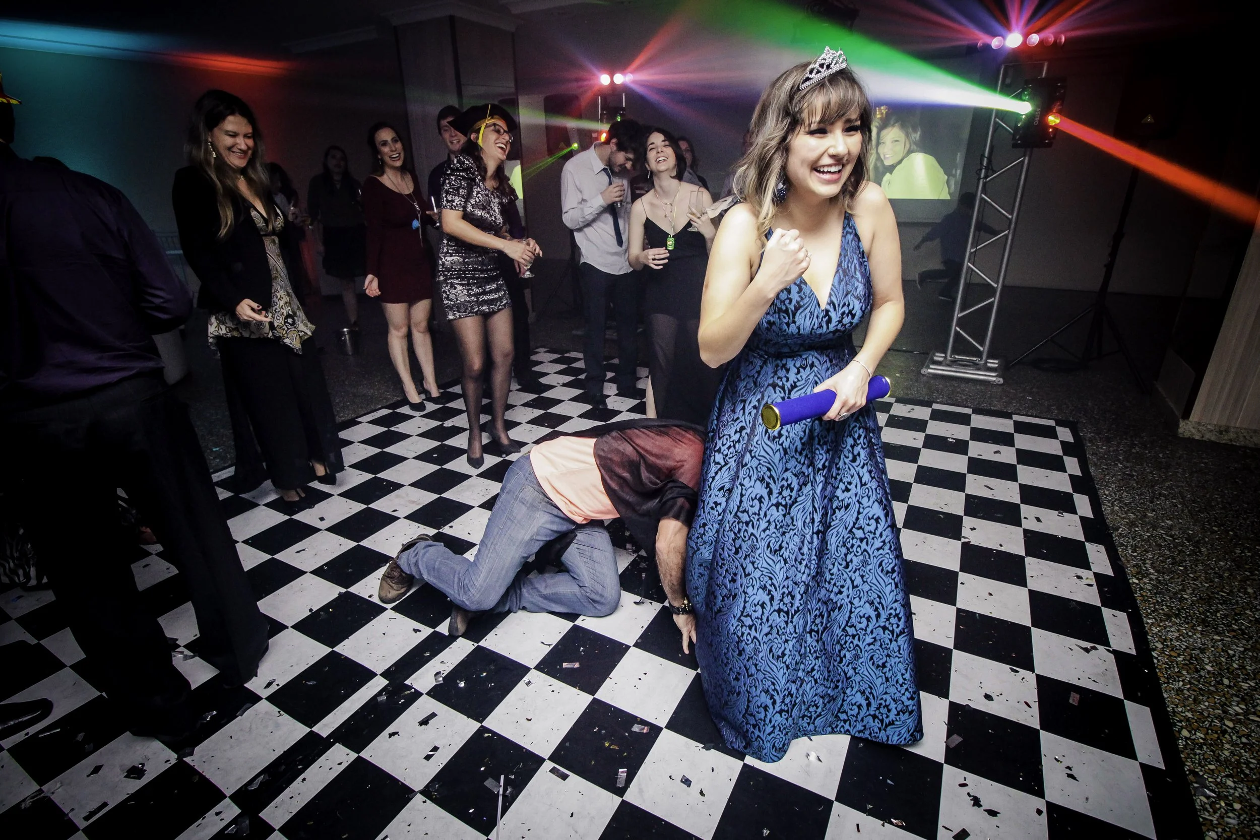 Mulher com tiara, sorrindo, segurando um monumento de Diploma no casamento, em uma festa com convidados ao fundo, na pista de dança com piso quadriculado.