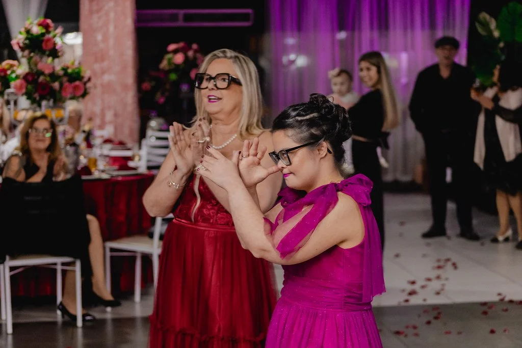 Duas mulheres dançando em uma celebração, uma de cabelo grisalho e outra com cabelo escuro, todas usando vestidos coloridos e óculos, com convidados ao fundo.