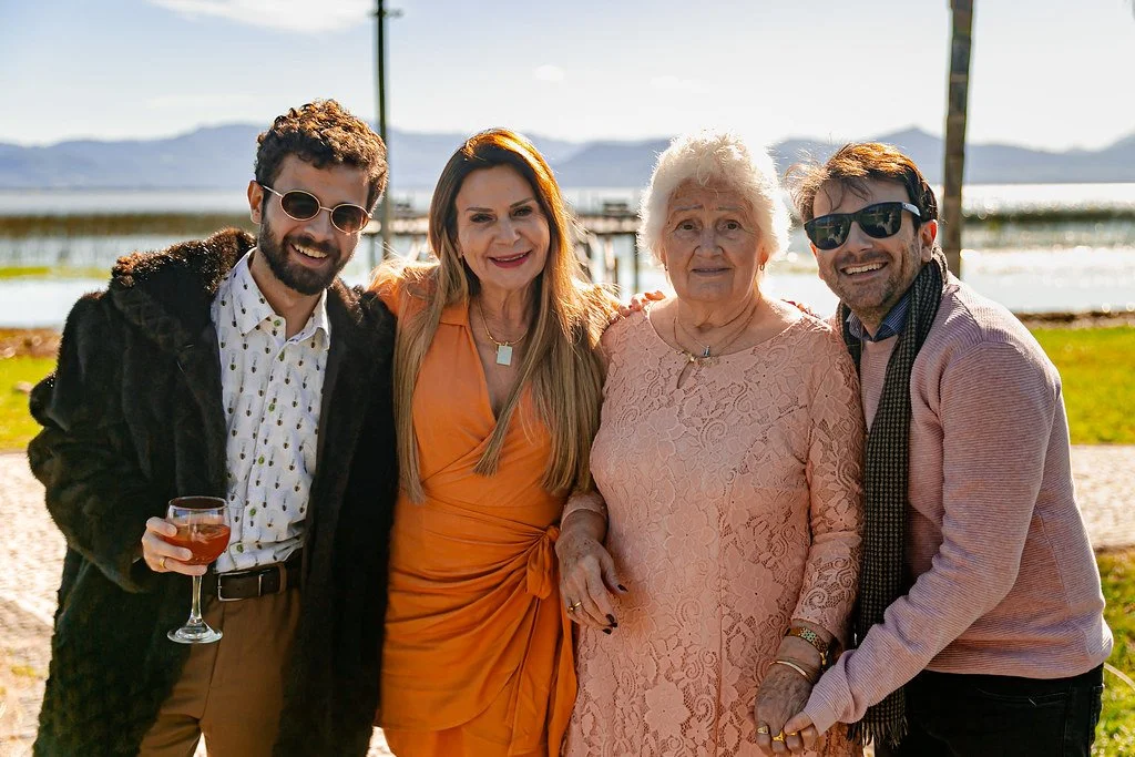 Quatro pessoas sorridentes posando ao ar livre, incluindo uma mulher idosa, um homem com óculos escuros, uma mulher jovem com vestido laranja, e um homem jovem com óculos escuros, ao fundo uma paisagem com lago e montanhas.
