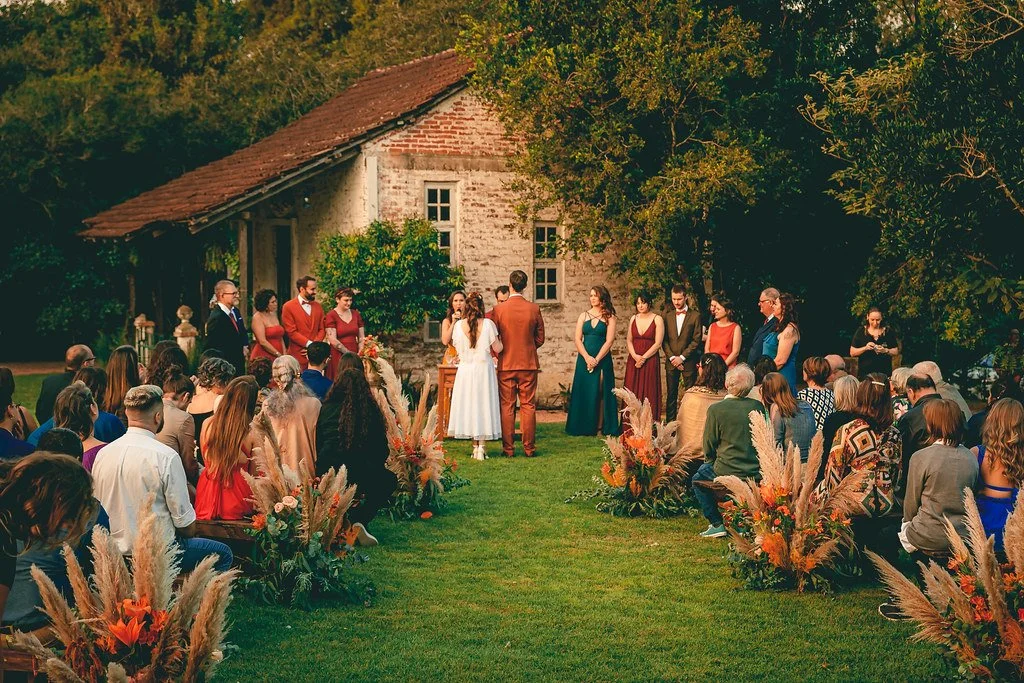 Casamento ao ar livre com noivos, padrinhos e convidados em frente a uma casa de campo, decorada com arranjos florais e grama verde
