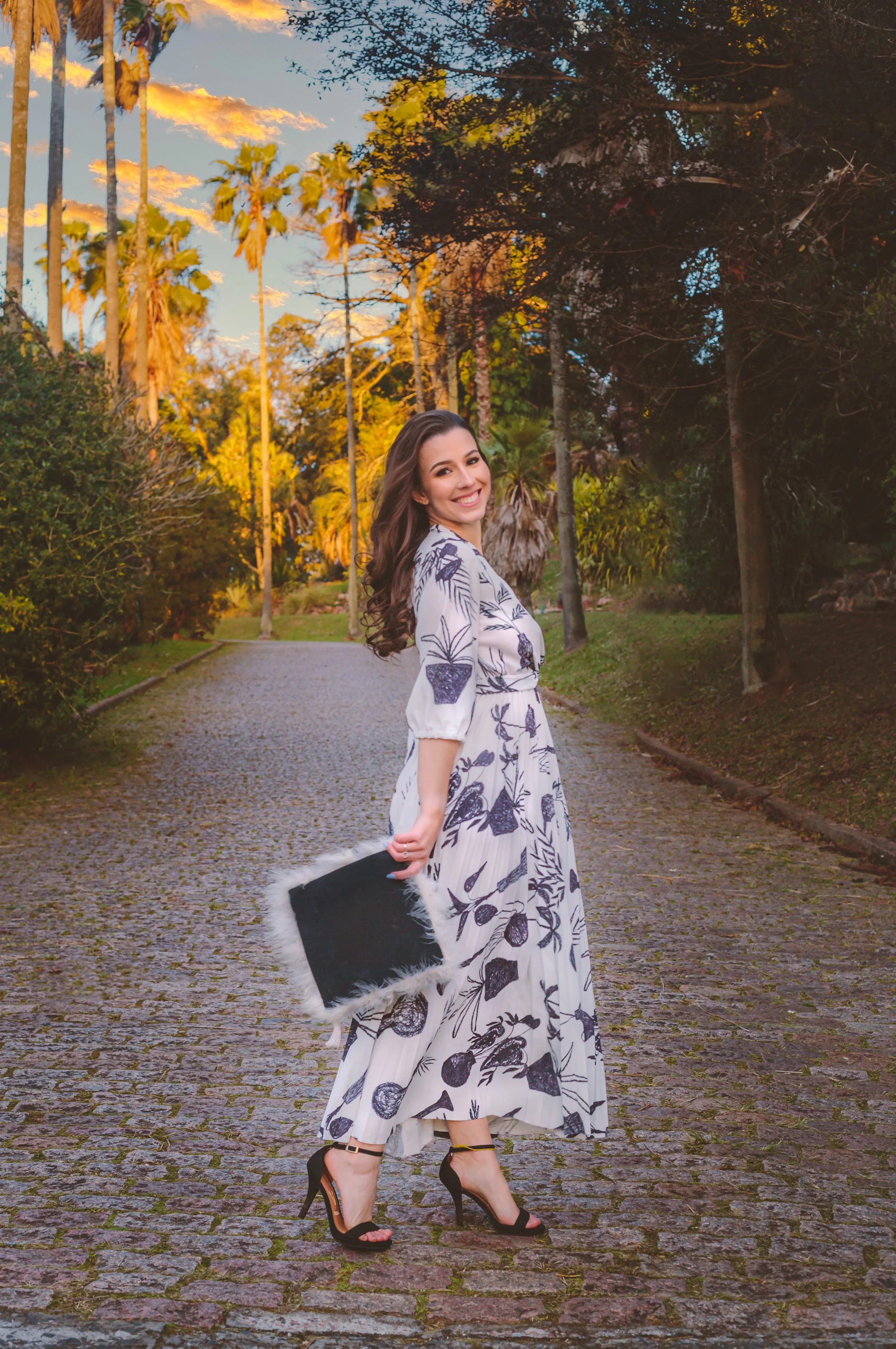 Mulher sorridente usando vestido longo com estampa de folhas, segurando uma pasta com detalhes de pele branca na borda, caminhando por um caminho de pedra em um parque com árvores, ao pôr do sol.