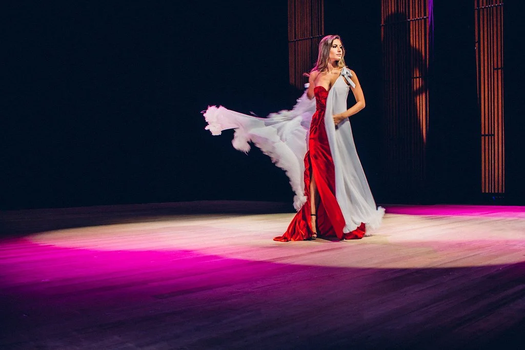 Mulher dançando no palco, vestindo um vestido longo branco e vermelho, com um vestido de festa de um ombro só, com iluminação em cores quentes.