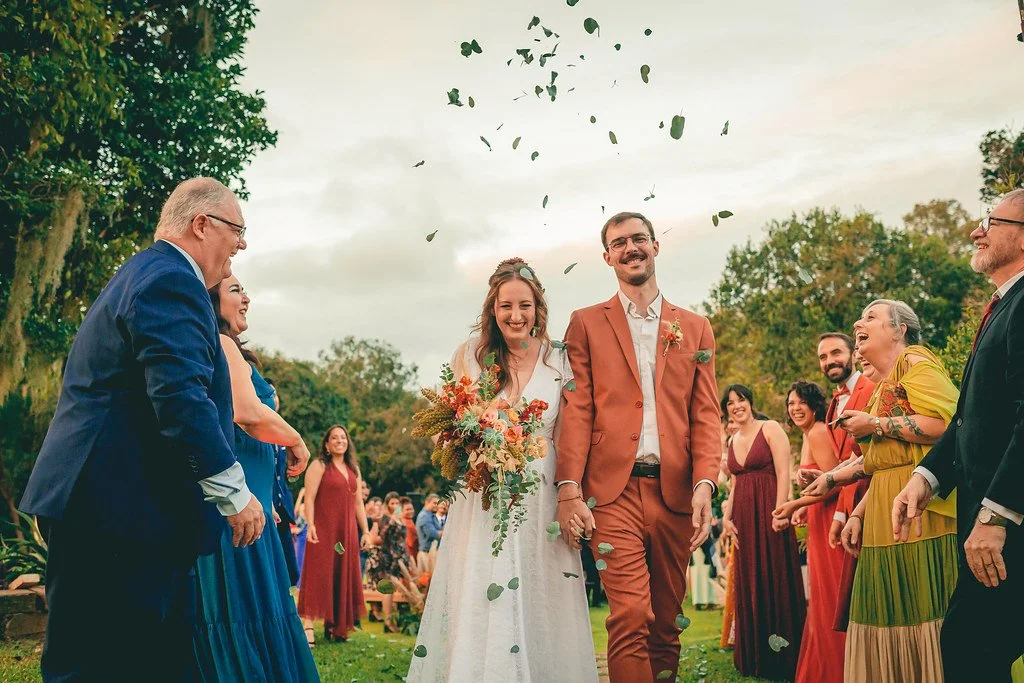 Casal de noivos caminhando ao ar livre, cercado por convidados jogando folhas, em um casamento ao ar livre, com árvores verdes ao fundo.