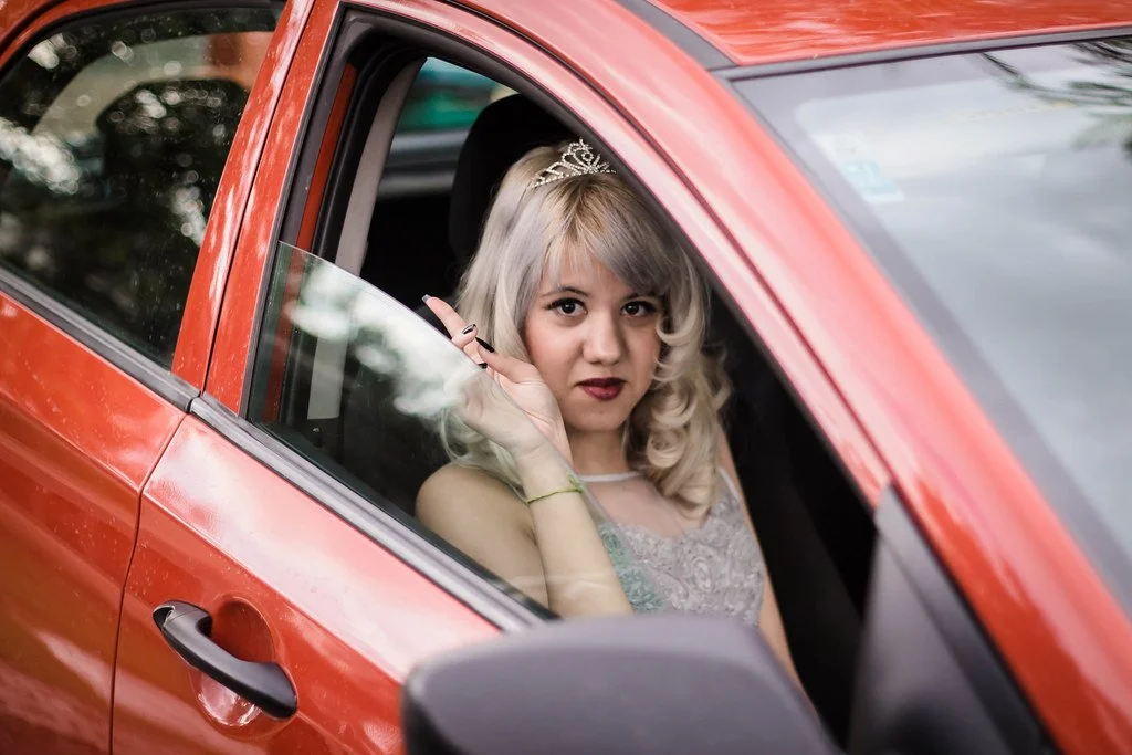 mulher com cabelo loiro encarando pela janela de um carro vermelho, usando uma tiara e vestido de festa.