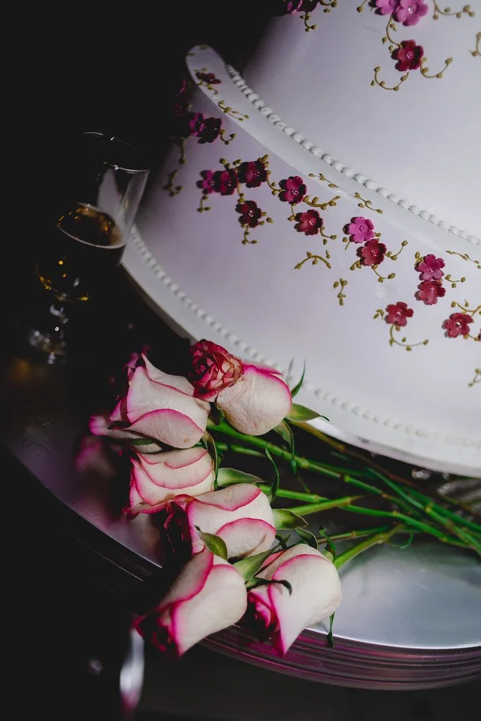 Bolo branco decorado com detalhes de flores e uma faixa de pérolas, acompanhado por rosas brancas com bordas rosa e um copo de bebida ao lado.
