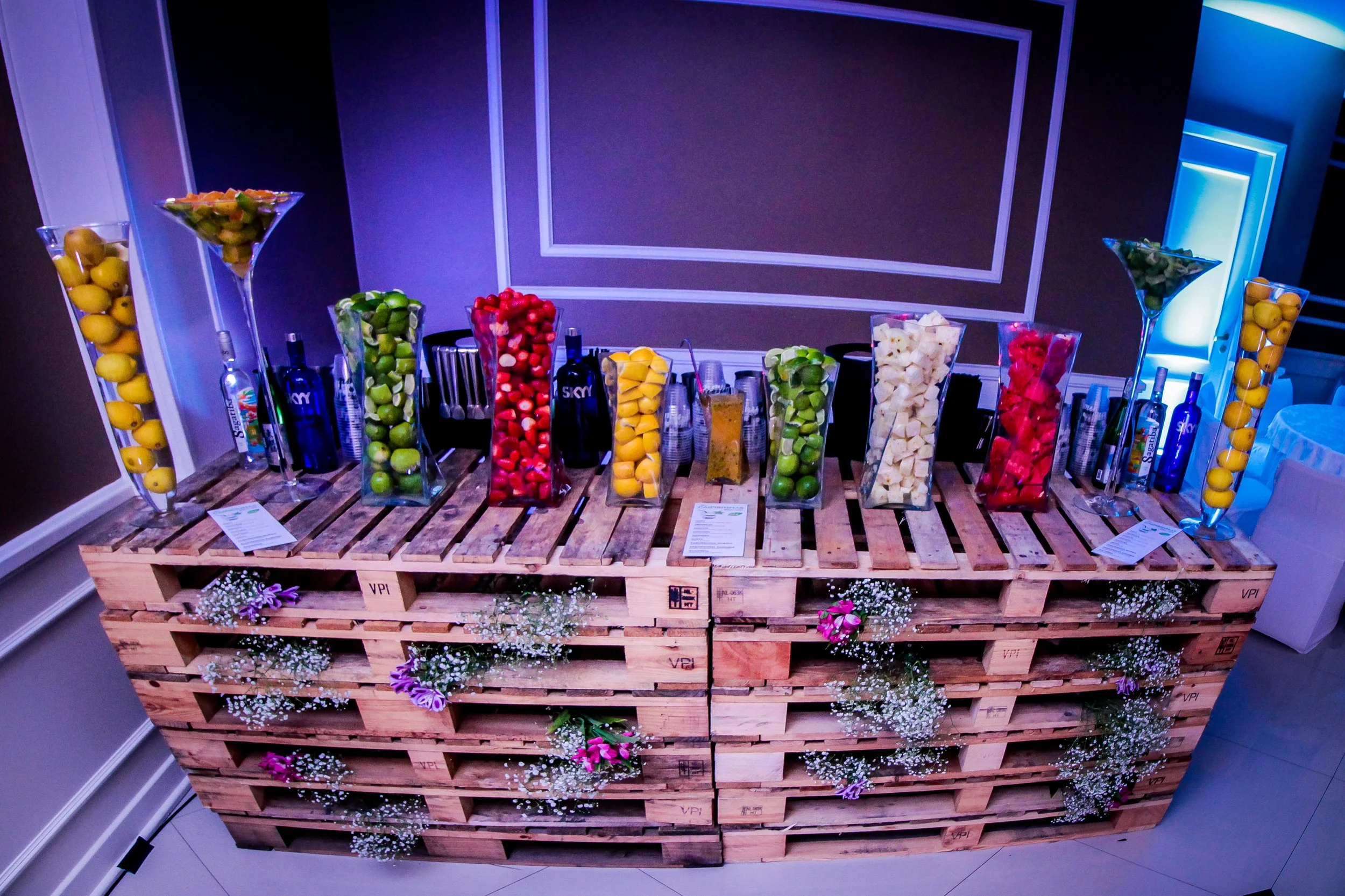Mesa decorada com garrafas de pallets de madeira, contendo frutas e bebidas coloridas em potes altos e garrafas, decorada com pequenas flores e cercada por um ambiente de festa.