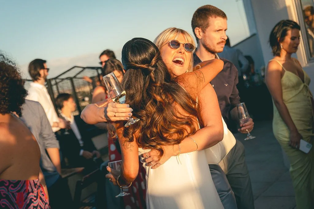 Mulher sorridente com óculos de sol abraçando uma outra pessoa, segurando uma taça de espumante, em uma festa ao ar livre com várias pessoas ao fundo.