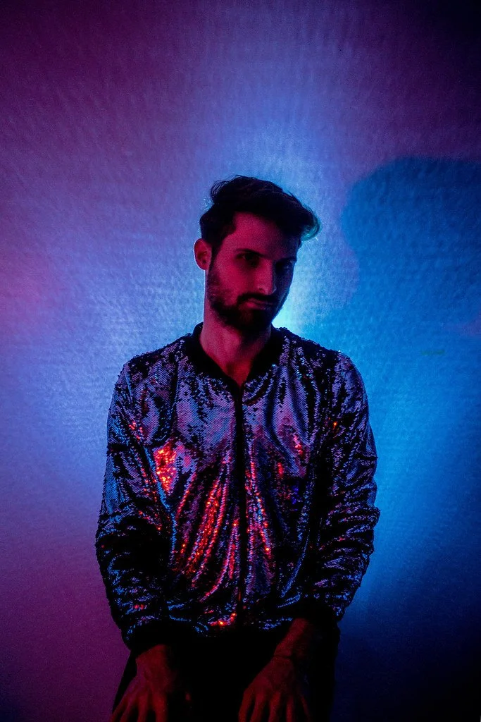 Homem de cabelo escuro e barba, usando jaqueta de paetês prateados, posando contra uma parede iluminada com luz de cores azuis e rosas.