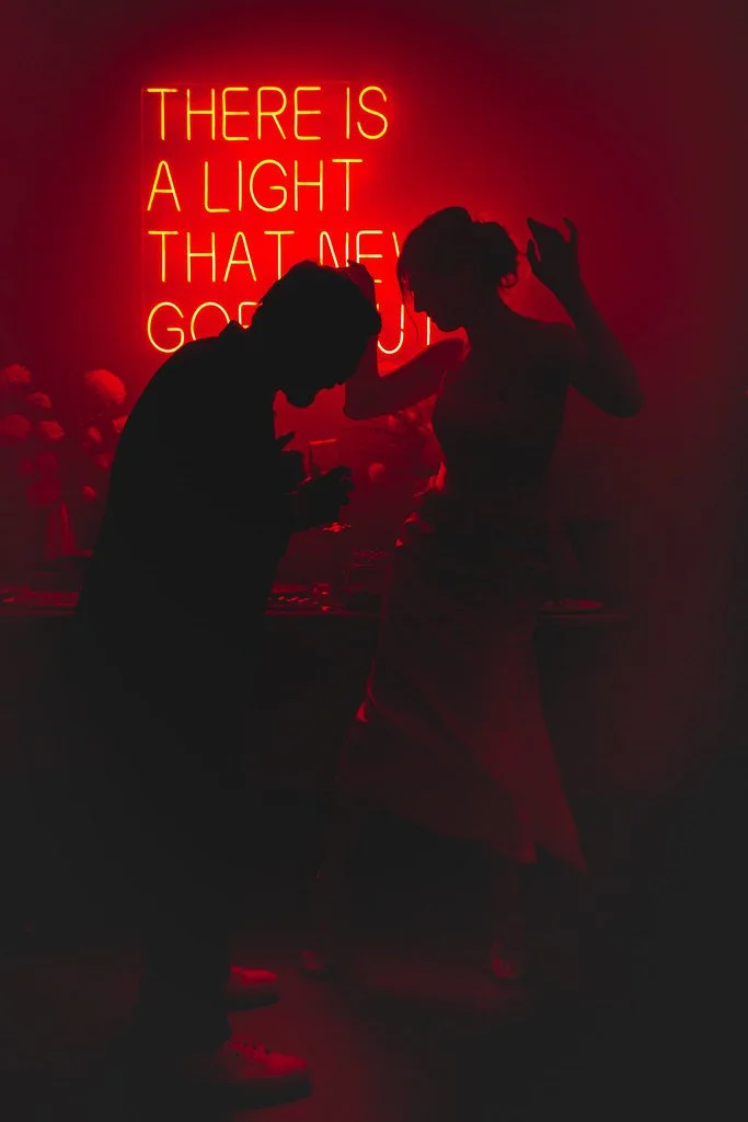 Silhuetas de um homem e uma mulher dançando sob iluminação de neon vermelha com a frase 'There is a light that never goes out' ao fundo.