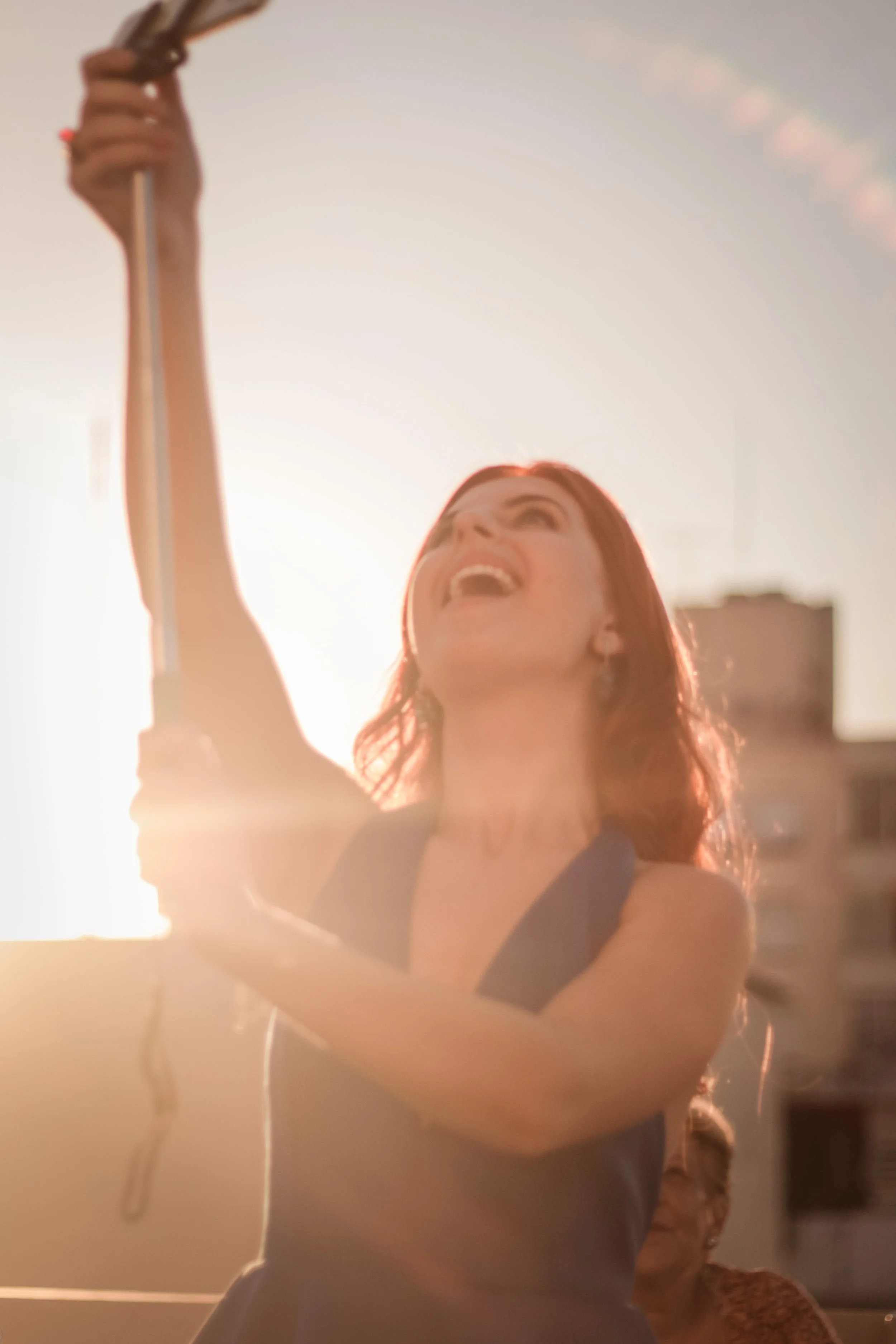 Mulher com cabelo vermelho, sorrindo e levantando o braço ao pôr do sol, em ambiente ao ar livre.