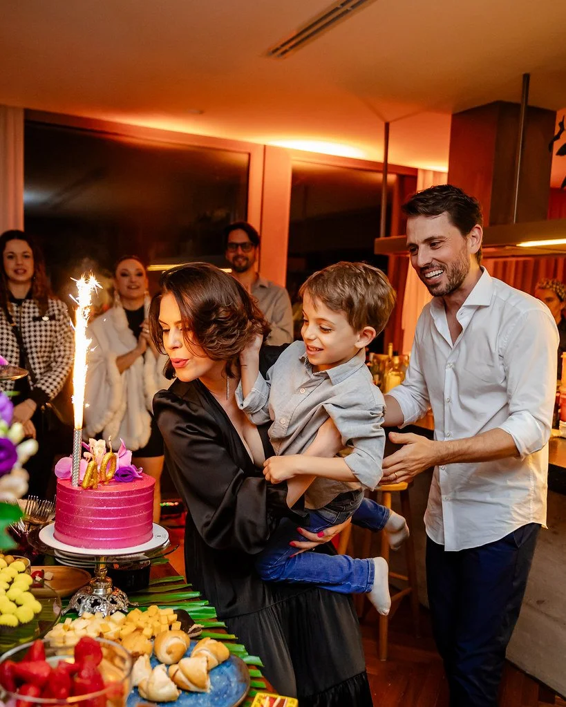 Família comemorando aniversário com bolo, brindando com festa de fogos de artifício e pessoas sorrindo ao fundo.