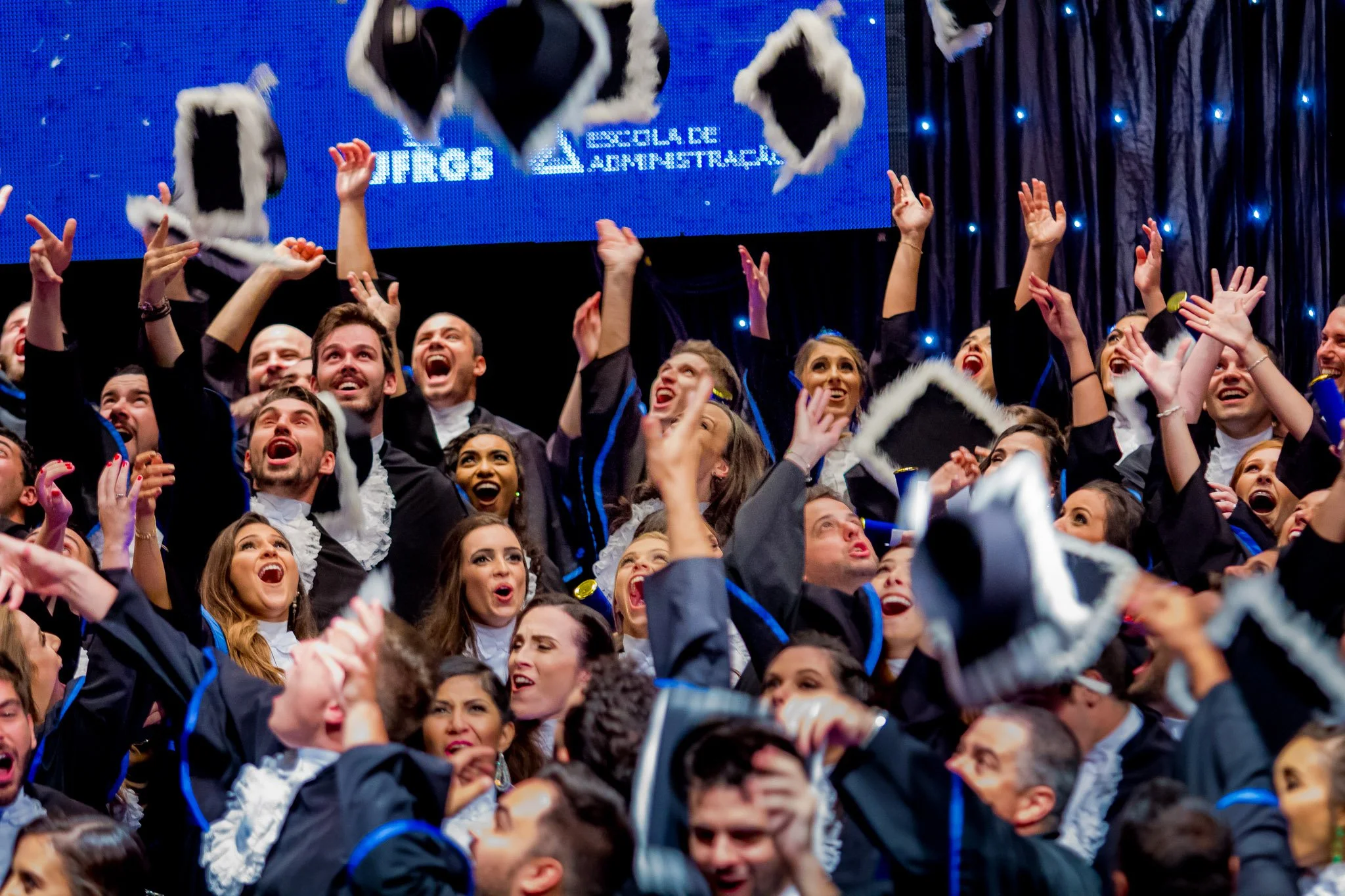 Celebration de formatura com estudantes lançando seus chapéus para o alto, sorrindo e comemorando em palco durante cerimônia de graduação