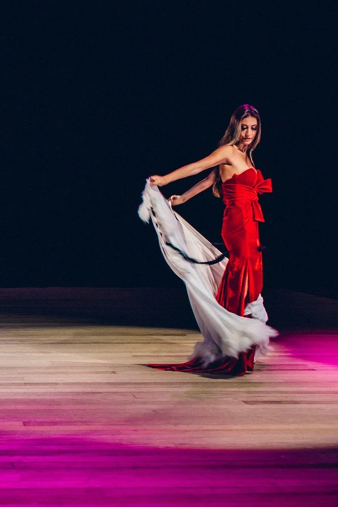 Mulher vestida com vestido vermelho em uma apresentação de dança, segurando uma saia branca com detalhes pretos, dançando em um palco de madeira com iluminação rosa.