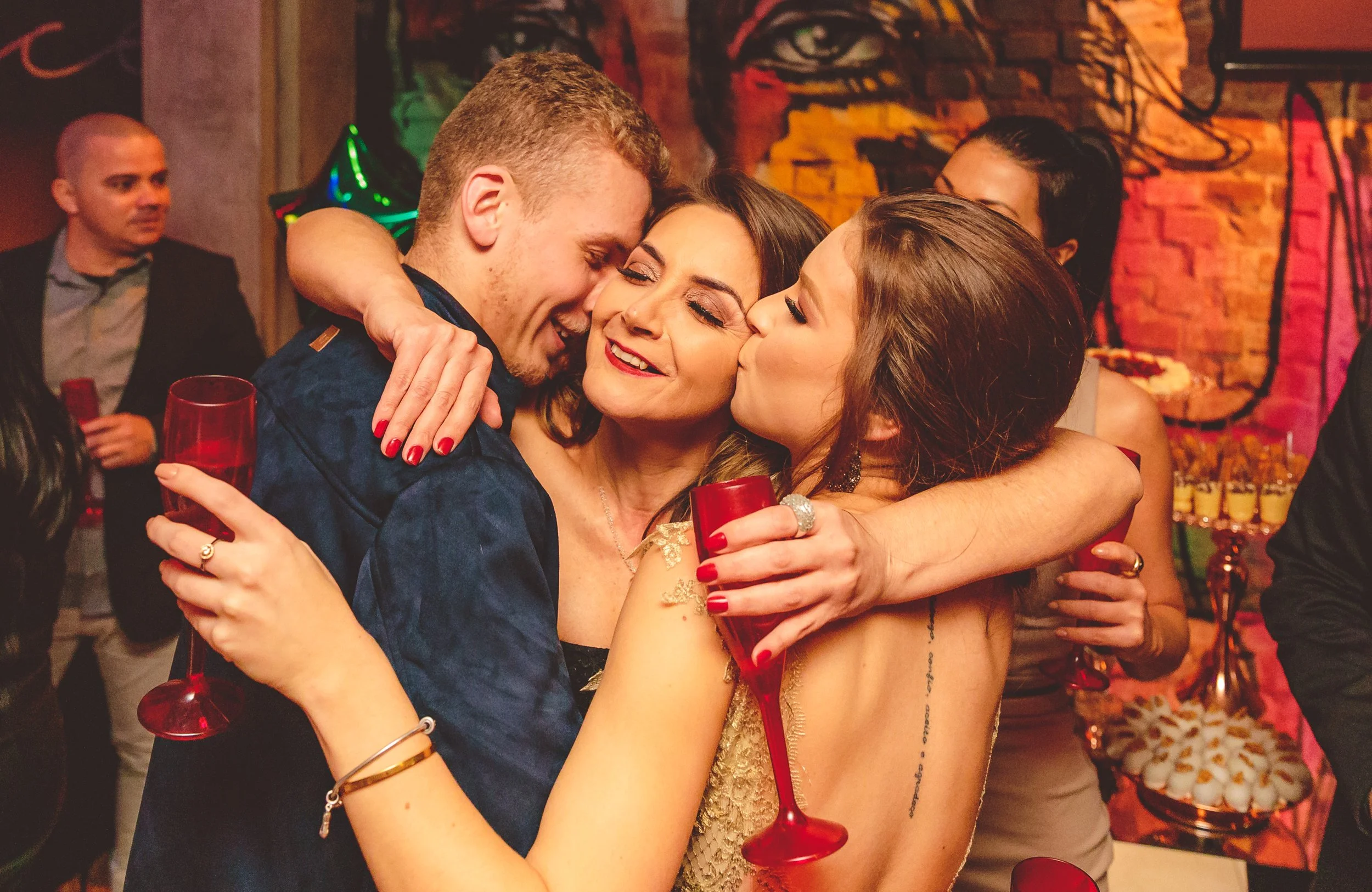 Pessoas se abraçando e sorrindo em uma festa, segurando taças de vinho vermelha.