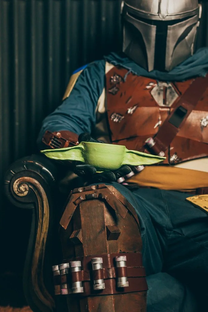 Personagem do universo Star Wars, usando capacete icônico, sentado em um sofá de madeira, segurando uma xícara de chá ou café. Bolinhos decorativos de aparência futurista na mão.