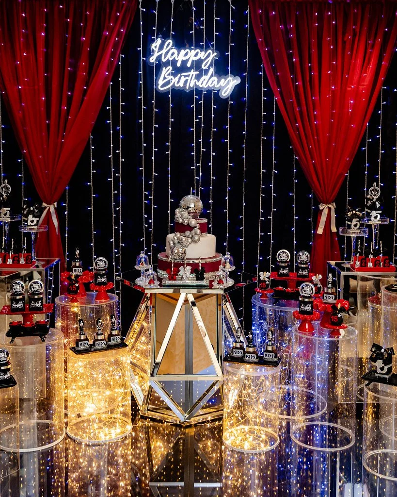 Decoração de aniversário com bolo central decorado com motivos prateados e vermelhos, luzes de pisca-pisca, e um letreiro luminoso que diz "Happy Birthday". Ao redor, mesas com lembrancinhas e elementos de festa, cortinas vermelhas com luzes azuis ao