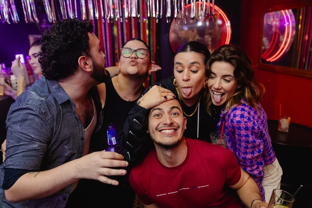 Grupo de cinco amigos se divertindo em um bar, brincando e sorrindo para a câmera.