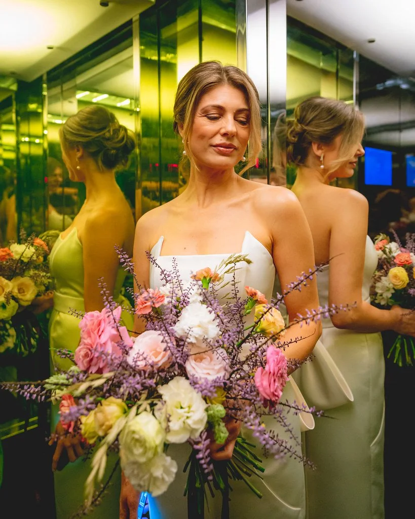 Mulher vestida com vestido branco, segurando um buquê de flores coloridas, em um ambiente com espelhos e iluminação amarela.