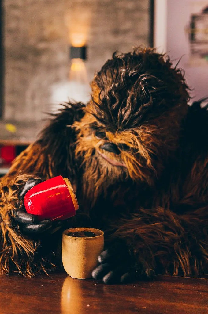 Cachorro de pelagem espessa e escura segurando uma xícara vermelha de café e uma xícara de cerâmica em uma mesa de madeira, ambiente aconchegante.