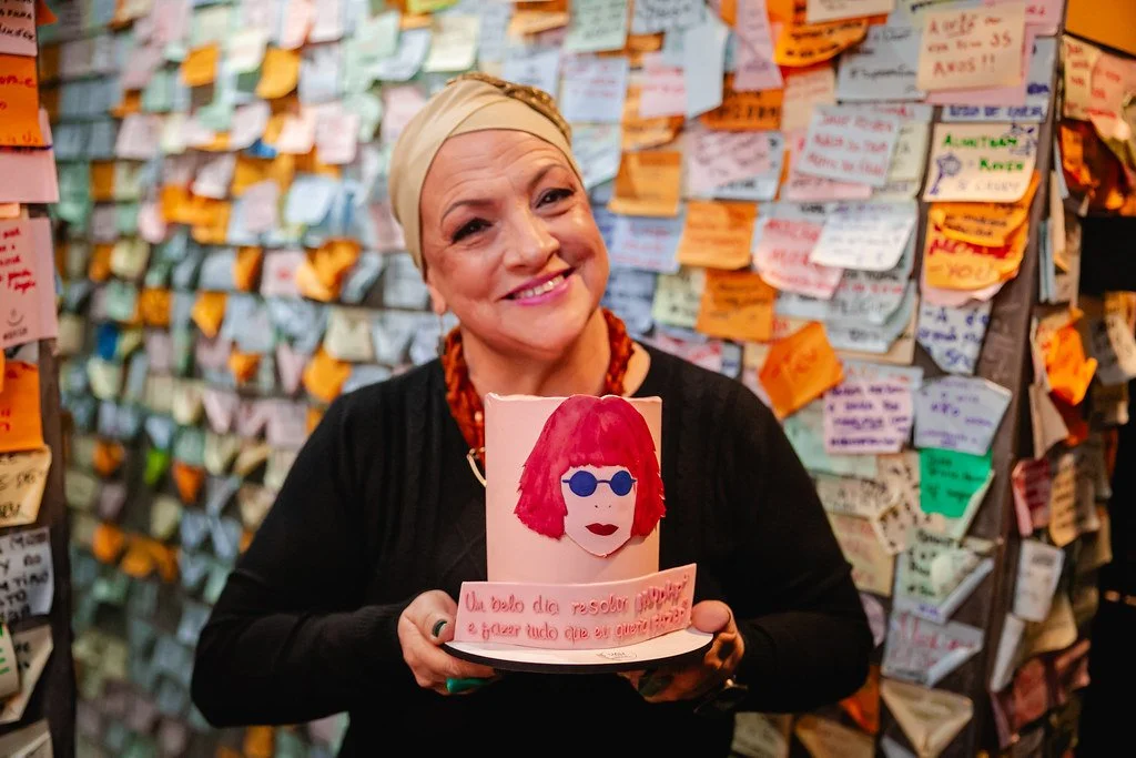 Mulher sorrindo segurando um cartaz com ilustração de uma pessoa de cabelo vermelho, óculos azuis e lábios vermelhos, no fundo cheio de papéis coloridos colados na parede.