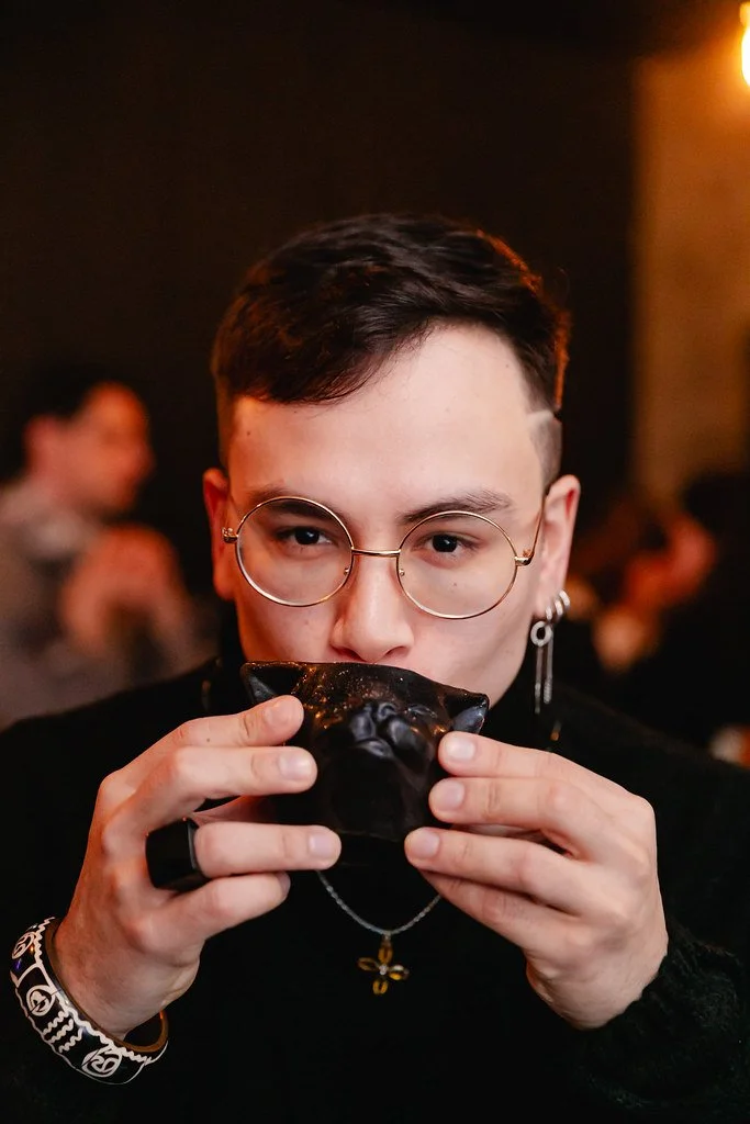 Pessoa com óculos, brincos e acessórios, segurando uma tigela preta próxima à boca, em ambiente com iluminação suave, com outras pessoas ao fundo.