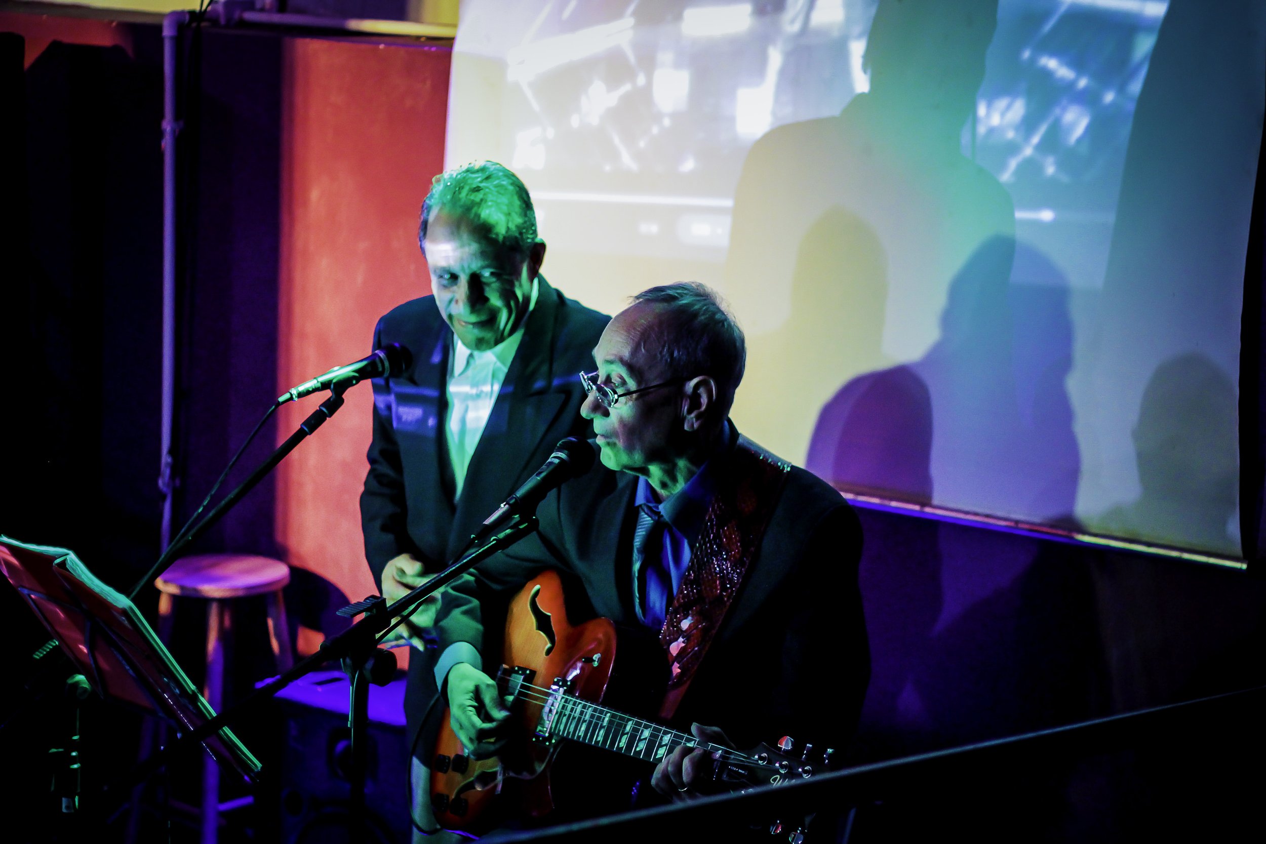 Dois homens em um palco, um tocando violão e outro cantando, com iluminação colorida