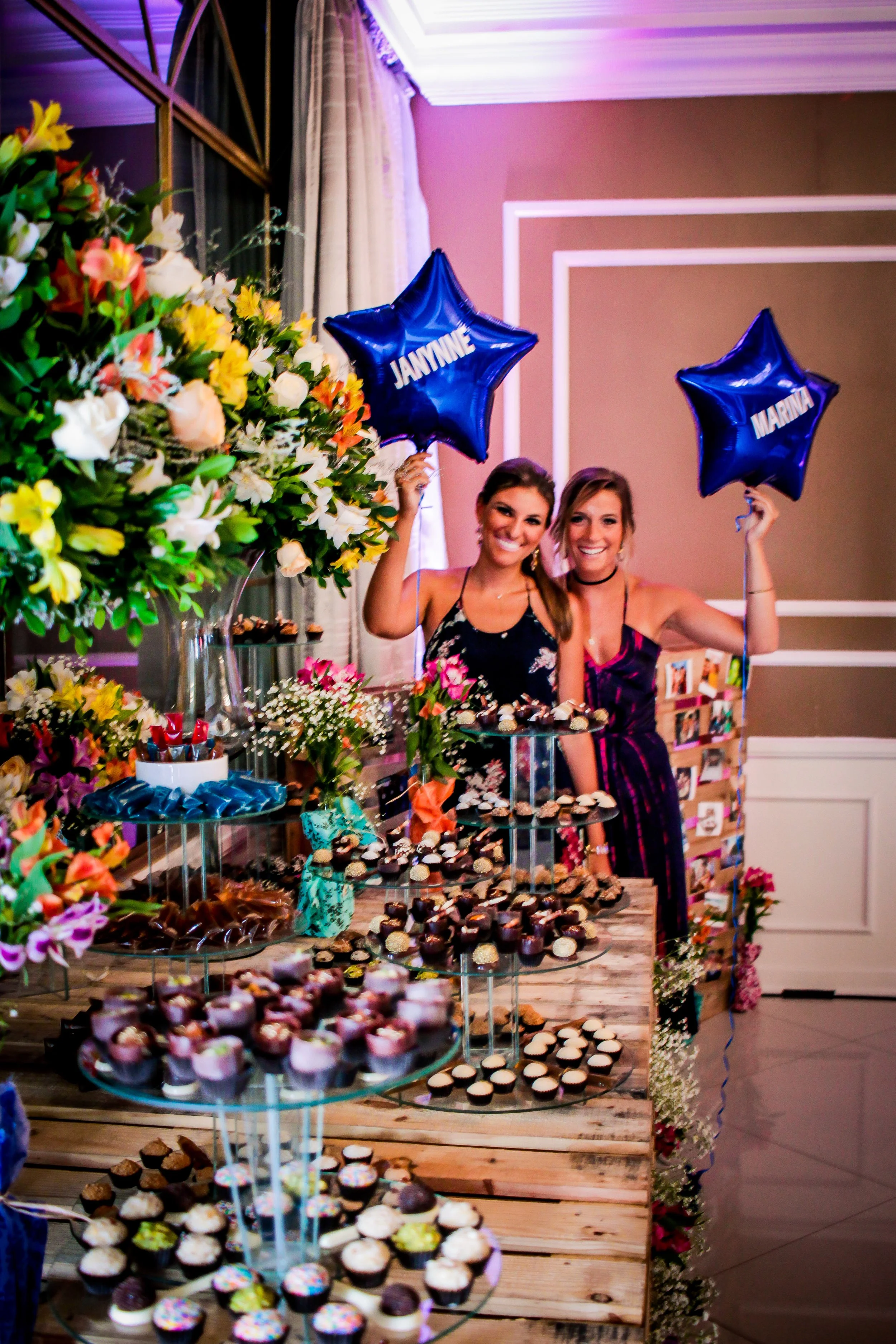 Duas meninas sorridentes comemorando em uma festa de aniversário com mesas de doces e balões azuis de estrelas escritos 'JAWNNE' e 'MARINA'.