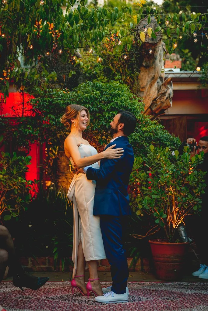Casal sorridente dançando ao ar livre, com decoração de luzes e plantas ao redor, durante evento social ou festa.