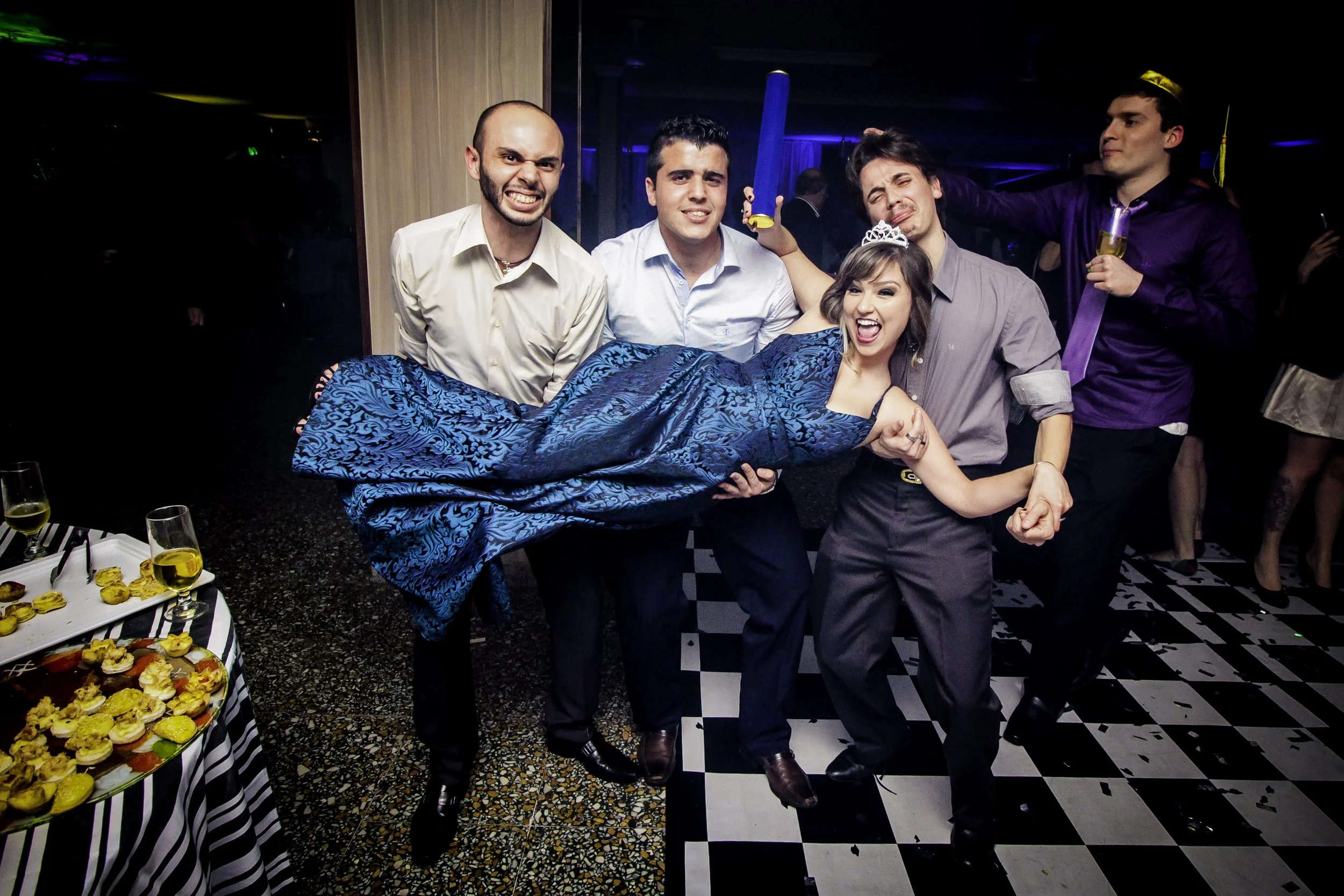 Grupo de pessoas celebrando festa, com uma mulher de vestido azul sendo carregada por cinco homens, alguns com expressões divertidas, em ambiente de festa com decoração e comida na mesa.
