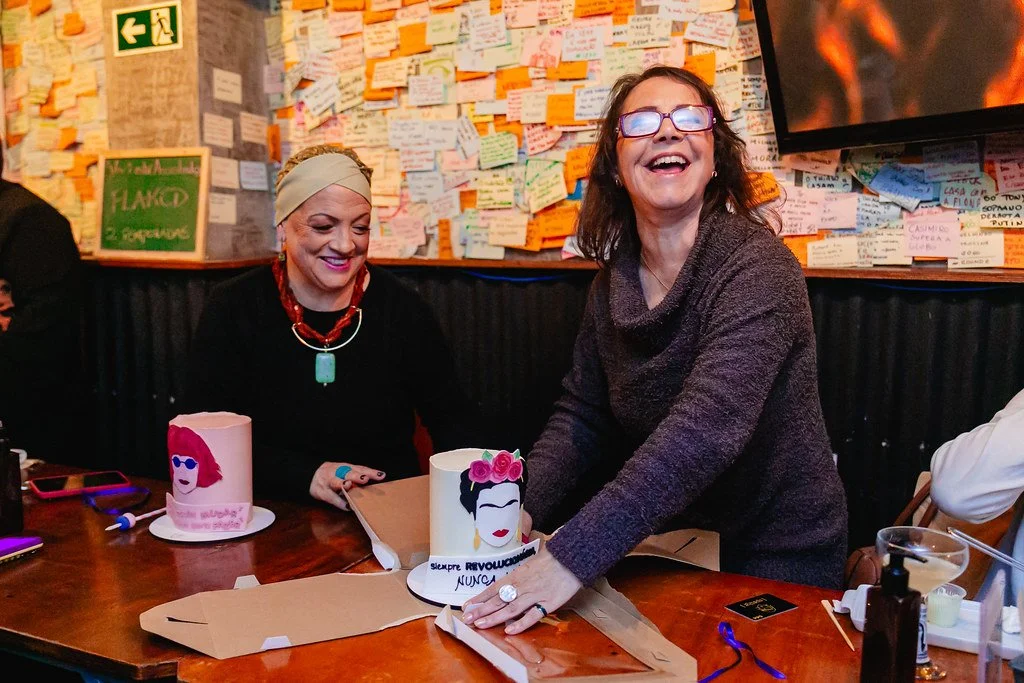 Duas mulheres sorridentes celebrando, uma segurando um bolo decorado com uma figura feminina e uma flor, em um ambiente com parede cheia de notas adesivas coloridas.