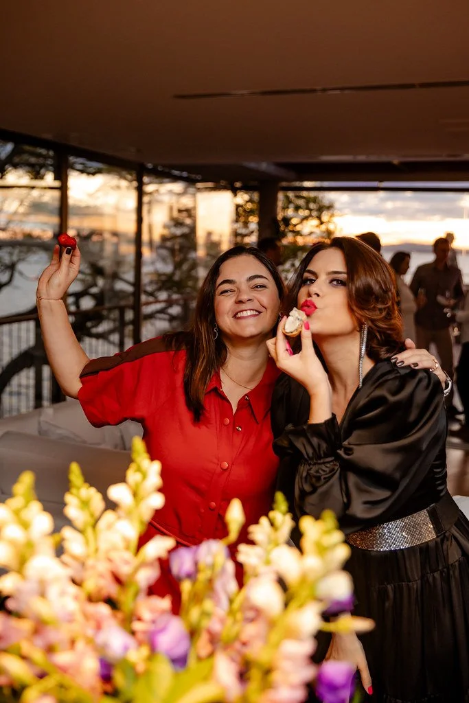Duas mulheres celebrando, uma com rosto sorridente e a outra fazendo um gesto de beijinho enquanto segura uma fatia de bolo, com um grupo de pessoas ao fundo e vista para um pôr do sol.