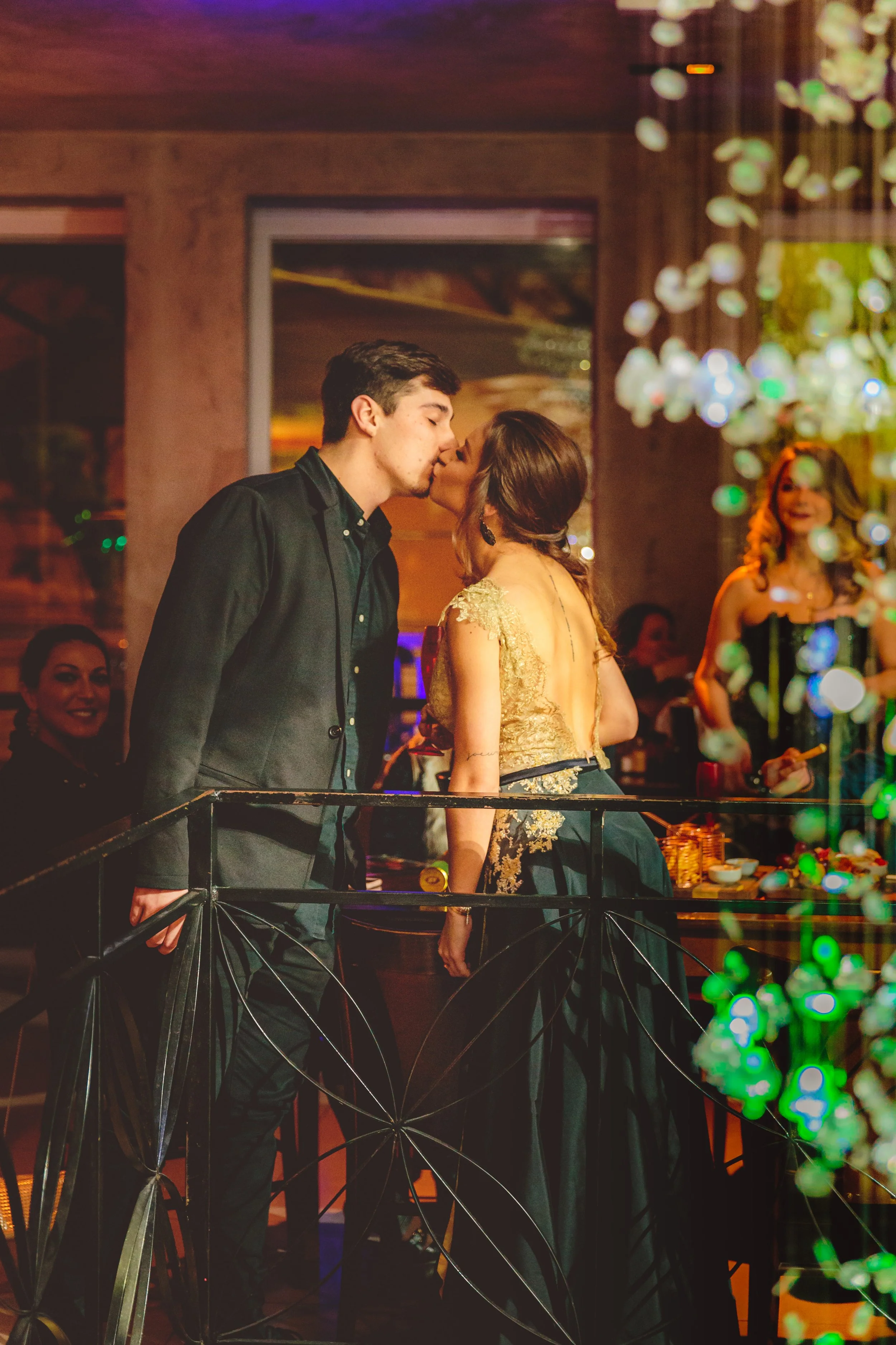 Um casal se beijando em uma festa, com várias pessoas ao fundo e decorações de luzes e plantas.