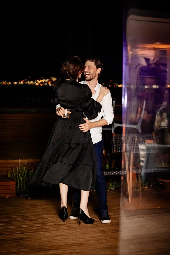 Casal alegre dançando de noite ao ar livre, com cidade iluminada ao fundo.